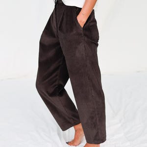 Könnte beinhalten: Eine Person trägt ein weißes T-Shirt, eine braune Cordhose und einen beigen Pullover über den Schultern. Die Hose hat eine hohe Taille und ein weites Bein. Das Outfit ist lässig und bequem.