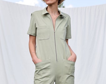 May include: A light green jumpsuit with short sleeves, a collared neckline, and a full-length zipper. The jumpsuit features two chest pockets and two large front pockets. The model is standing with their hands in the pockets.
