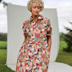 May include: A floral print dress with short sleeves and a button-down front. The dress features a collared neckline and a gathered waist. The fabric has a pattern of red, orange, blue, and green flowers. The dress is knee-length and has pockets.