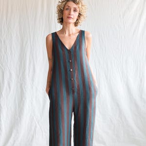 Op de afbeelding: Een mouwloze, gestreepte jumpsuit in teal en bruin. De jumpsuit met wijde pijpen heeft een V-hals, een knoopsluiting aan de voorkant en zakken. Het model draagt de jumpsuit voor een witte achtergrond.