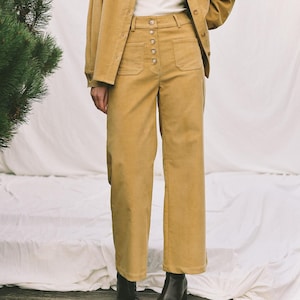Könnte beinhalten: Senfgelbes Cord-Ensemble, bestehend aus einer Jacke mit Knopfleiste und einer passenden hoch taillierten Hose. Die Jacke hat eine Brusttasche. Die Hose hat große Vordertaschen und einen Knopfverschluss. Das Model trägt ein weißes T-Shirt und schwarze Split-Toe-Stiefel.