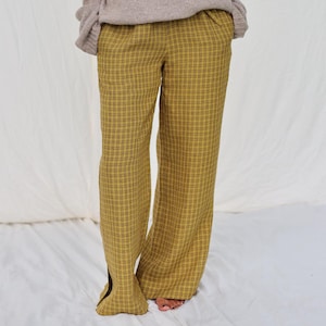 Puede incluir: Una persona con un suéter beige de cuello redondo y pantalones de cuadros amarillos. Los pantalones tienen una pernera ancha y una abertura en el dobladillo. El atuendo es informal y cómodo.