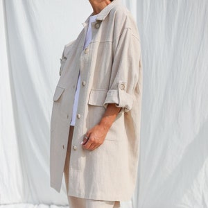 May include: A beige linen jacket with a button-up front and two large pockets. The jacket is worn over a white t-shirt and beige linen pants. The person is wearing blue suede clogs.
