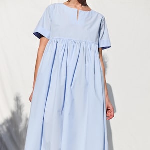 Può includere: Un abito midi azzurro con maniche corte, scollo rotondo con un piccolo spacco verticale e vita arricciata che crea una gonna ampia e fluida. L'abito è abbinato a scarpe da ginnastica bianche stringate con suola in gomma color marrone chiaro.