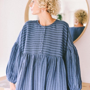 May include: A woman wearing a blue and white striped dress with a button-up front. The dress has a loose fit and long sleeves.