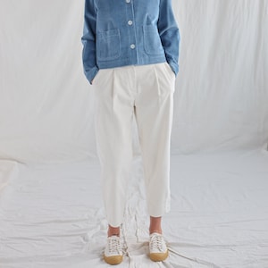 May include: A woman wearing a light blue corduroy jacket with a white collar and white pants. She is standing in front of a white background.