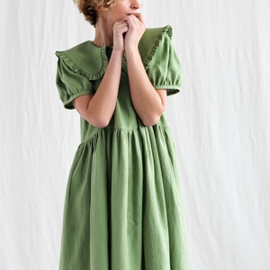 Può includere: Una donna indossa un vestito di lino verde con un colletto a volant e maniche corte. È in piedi su uno sfondo bianco e guarda di lato. Indossa scarpe da ginnastica grigie.