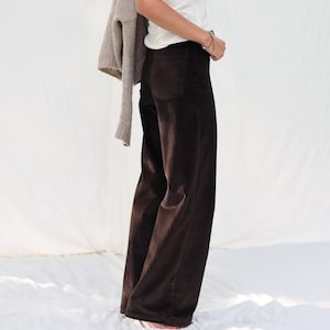 Könnte beinhalten: Eine Frau trägt ein weißes Tanktop und eine dunkelbraune Cordhose. Ein hellbrauner Pullover ist über den Schultern drapiert. Die Hose hat ein weites Bein-Design.