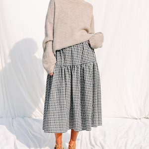 May include: A woman is wearing a beige sweater and a black and white gingham skirt. She is wearing brown shoes.