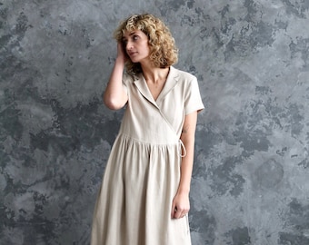 May include: A woman wearing a beige linen wrap dress with short sleeves and a collared neckline. She is standing in front of a textured gray wall and wearing black sandals.
