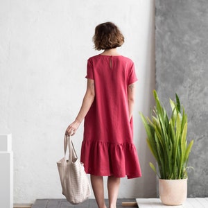 May include: A red linen dress with short sleeves and a ruffled hem. The person is holding a striped tote bag and wearing silver shoes. A potted plant is in the background.
