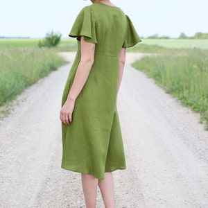 Può includere: Una donna indossa un abito di lino verde con maniche corte e arricciate. È in piedi su una strada sterrata con erba verde su entrambi i lati.