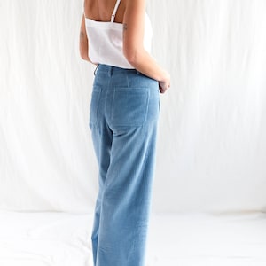 May include: A white camisole with thin shoulder straps paired with light blue corduroy trousers featuring a high waist, belt loops, and two large patch pockets on the back. The trousers have a straight-leg cut and a vertical ribbed texture.