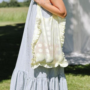 May include: A light blue and white striped sleeveless dress with a tiered skirt and a pale yellow tote bag featuring a ruffled trim and a subtle white floral pattern. The dress has a button-down front and a loose, flowing silhouette.