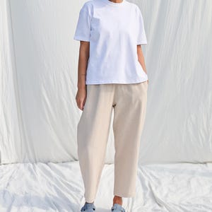 Puede incluir: Una camiseta blanca de gran tama&ntilde;o con mangas cortas y un corte holgado combinada con pantalones beige de pierna ancha. La modelo lleva zapatos planos de color azul claro.