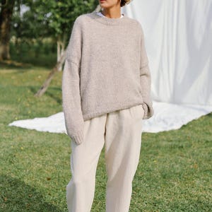Puede incluir: Una mujer con un su&eacute;ter de lana marr&oacute;n claro y pantalones beige. Est&aacute; de pie en un c&eacute;sped verde con una s&aacute;bana blanca detr&aacute;s de ella.