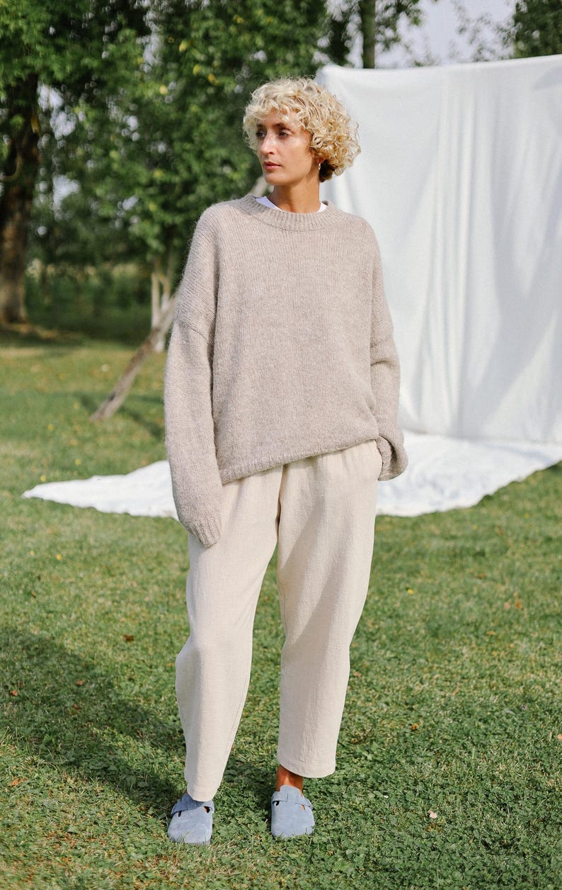 Puede incluir: Una mujer con un su&eacute;ter de lana marr&oacute;n claro y pantalones beige. Est&aacute; de pie en un c&eacute;sped verde con una s&aacute;bana blanca detr&aacute;s de ella.