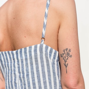 Puede incluir: Una mujer con un vestido de lino a rayas azules y blancas con una tira fina. Tiene un peque&ntilde;o tatuaje de flor en la parte superior del brazo.