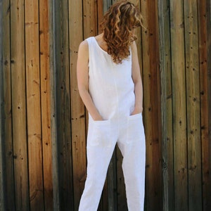 May include: A sleeveless white jumpsuit featuring a round neckline, two large square patch pockets on the front, and tapered pant legs with cuffed hems. The garment is paired with brown leather clogs that have a buckle strap and wooden soles.