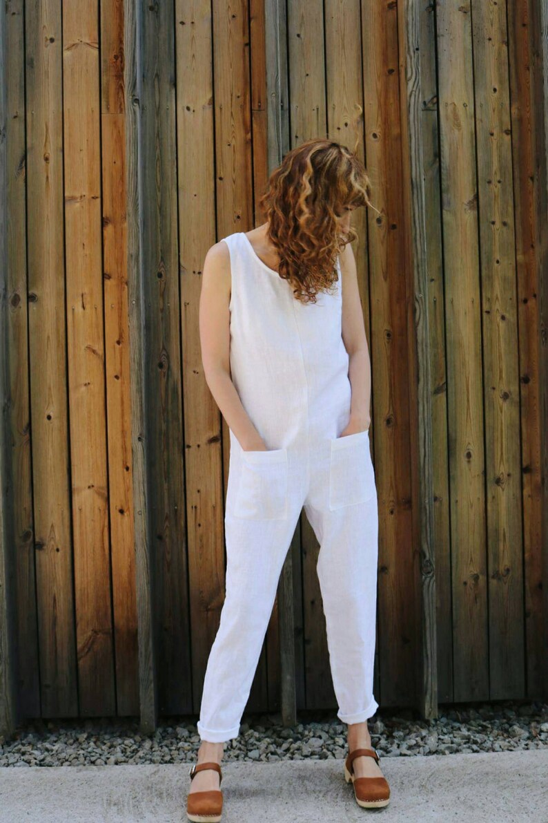 May include: A sleeveless white jumpsuit featuring a round neckline, two large square patch pockets on the front, and tapered pant legs with cuffed hems. The garment is paired with brown leather clogs that have a buckle strap and wooden soles.