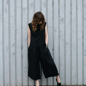 May include: A sleeveless black jumpsuit featuring a round neckline, a dropped waistline with a gathered skirt section, and wide-leg trousers. The garment includes side pockets and is paired with black lace-up shoes, creating a solid dark silhouette.