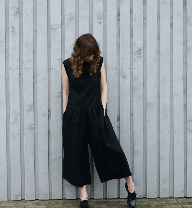 May include: A sleeveless black jumpsuit featuring a round neckline, a dropped waistline with a gathered skirt section, and wide-leg trousers. The garment includes side pockets and is paired with black lace-up shoes, creating a solid dark silhouette.