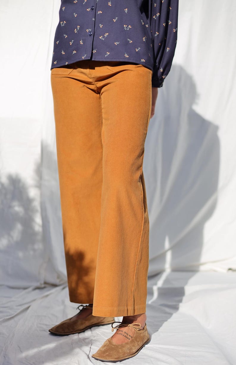 May include: A pair of wide-leg corduroy pants in a mustard yellow color. The pants have a straight leg and a relaxed fit. The model is wearing the pants with a navy blue floral print shirt and brown leather shoes.