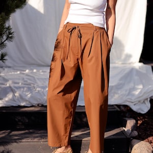 Puede incluir: Pantalón ancho marrón con cintura de cordón y pliegues. El pantalón está hecho de una tela ligera y tiene un corte holgado.
