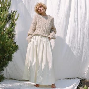 Puede incluir: Una mujer con un suéter beige de punto trenzado y una falda larga blanca. El suéter tiene cuello redondo y mangas largas. La falda es de cintura alta y llega hasta el suelo. El fondo es una sábana blanca.