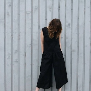 May include: A solid black sleeveless jumpsuit featuring a round neckline, a fitted bodice, and wide-leg cropped trousers. The garment includes side pockets and a seam at the waist, providing a clean, structured silhouette with a simple, uniform design.