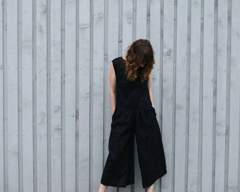 May include: A solid black sleeveless jumpsuit featuring a round neckline, a fitted bodice, and wide-leg cropped trousers. The garment includes side pockets and a seam at the waist, providing a clean, structured silhouette with a simple, uniform design.