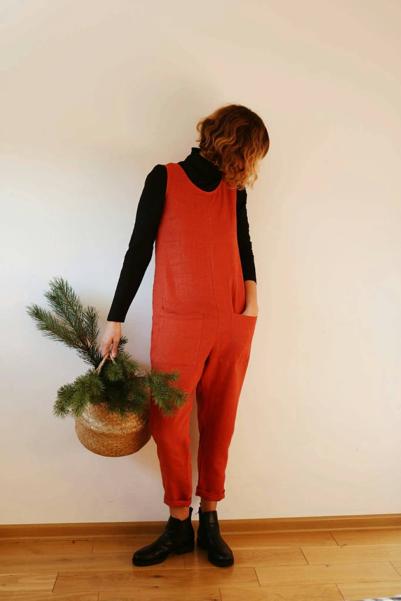 Puede incluir: Una mujer lleva un mono rojo con una camiseta de cuello alto negra y botines negros. Lleva una cesta de mimbre con una rama de pino.