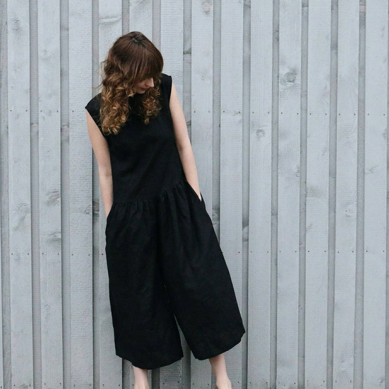May include: A sleeveless black jumpsuit featuring a round neckline, a fitted bodice, and a dropped waistline that transitions into wide-leg trousers. The garment includes side pockets and is constructed from a solid black fabric with a matte finish.