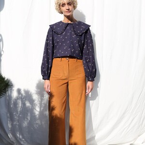 May include: A woman wearing a navy blue blouse with a white ruffled collar and a floral print. She is also wearing brown corduroy pants.