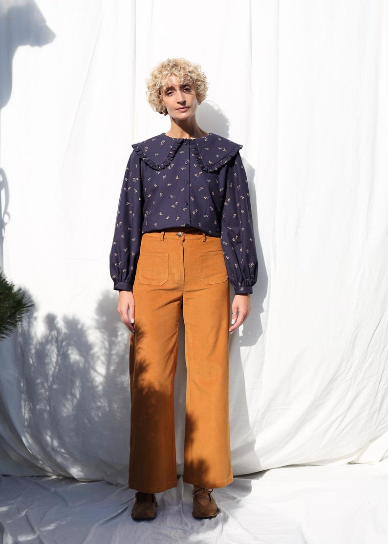 May include: A woman wearing a navy blue blouse with a white ruffled collar and a floral print. She is also wearing brown corduroy pants.