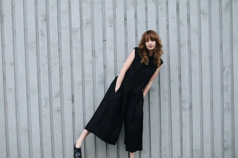 May include: A sleeveless black jumpsuit featuring a round neckline, a fitted bodice, and wide-leg trousers that fall below the knee. The garment includes two side pockets and is paired with black lace-up shoes, creating a functional and simple look.