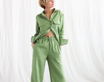 Puede incluir: Un conjunto de camisa y pantalón de lino verde. La camisa tiene un frente abotonado y mangas largas. El pantalón tiene una cintura con cordón y piernas anchas.