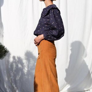 May include: A woman wearing a navy blue blouse with a white collar and a floral pattern. She is wearing mustard yellow wide-leg pants and brown shoes.