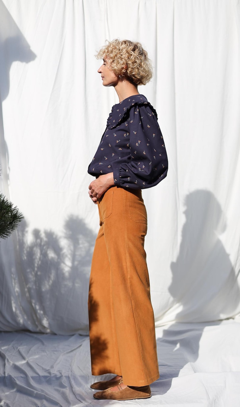 May include: A woman wearing a navy blue blouse with a white collar and a floral pattern. She is wearing mustard yellow wide-leg pants and brown shoes.