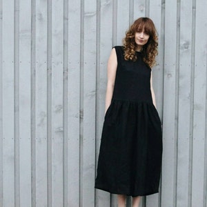 May include: A sleeveless black midi dress featuring a round neckline and a dropped waistline with a gathered, voluminous skirt. The garment is constructed with a fitted bodice that transitions into a relaxed, full skirt silhouette that falls below the knee.