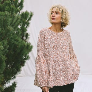 May include: A cream-coloured blouse with a floral pattern in shades of red and brown. The long-sleeved top has a round neckline and a tiered design. The sleeves are wide and flared. The model is wearing black trousers.