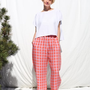 May include: A woman wearing a white linen top and pink and orange gingham pants. She is standing in front of a white backdrop.