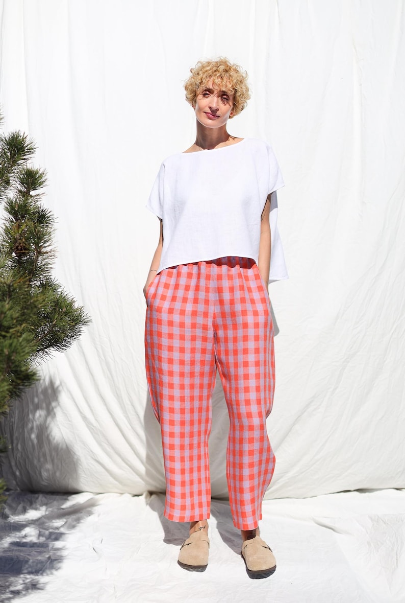 May include: A woman wearing a white linen top and pink and orange gingham pants. She is standing in front of a white backdrop.