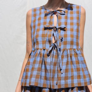May include: A sleeveless top and skirt set in a blue and brown plaid pattern. The top features a keyhole neckline with bow ties and a ruffled hem. The fabric has a soft, textured appearance, suggesting a lightweight material.