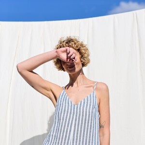 Puede incluir: Una mujer con el pelo rubio rizado lleva un vestido de lino a rayas azules y blancas. Est&aacute; de pie frente a un fondo blanco y se tapa un ojo con la mano.
