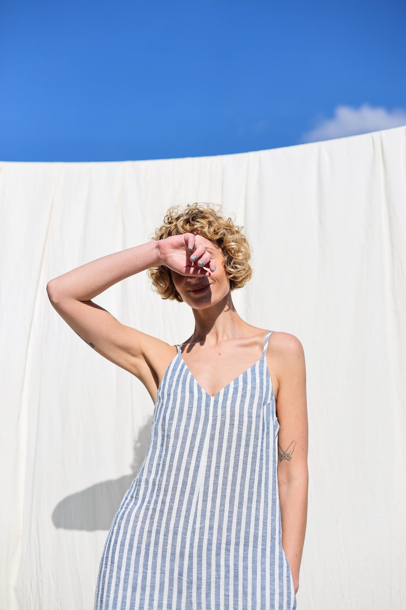 Puede incluir: Una mujer con el pelo rubio rizado lleva un vestido de lino a rayas azules y blancas. Est&aacute; de pie frente a un fondo blanco y se tapa un ojo con la mano.
