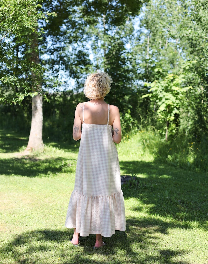 May include: A floor-length, sleeveless dress featuring a light beige and white gingham check pattern. The garment has thin shoulder straps, a straight neckline, and a wide, gathered ruffle hem that adds volume to the lower portion of the skirt.