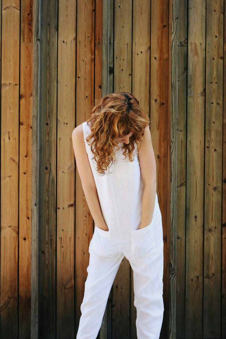 May include: A sleeveless white jumpsuit featuring a round neckline and two large patch pockets on the front. The garment has a relaxed fit with tapered legs and is constructed from a solid white fabric, providing a clean and simple aesthetic.