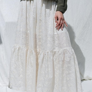 May include: A white, floor-length skirt with a floral embroidered pattern. The skirt has a tiered, ruffled hem. A dark green jacket is worn above the skirt. The image is taken in natural light.
