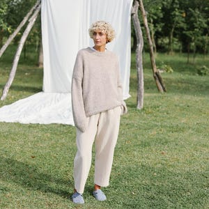 Puede incluir: Una mujer con un su&eacute;ter marr&oacute;n claro y pantalones blancos. Est&aacute; de pie en un &aacute;rea cubierta de hierba con una s&aacute;bana blanca detr&aacute;s de ella.